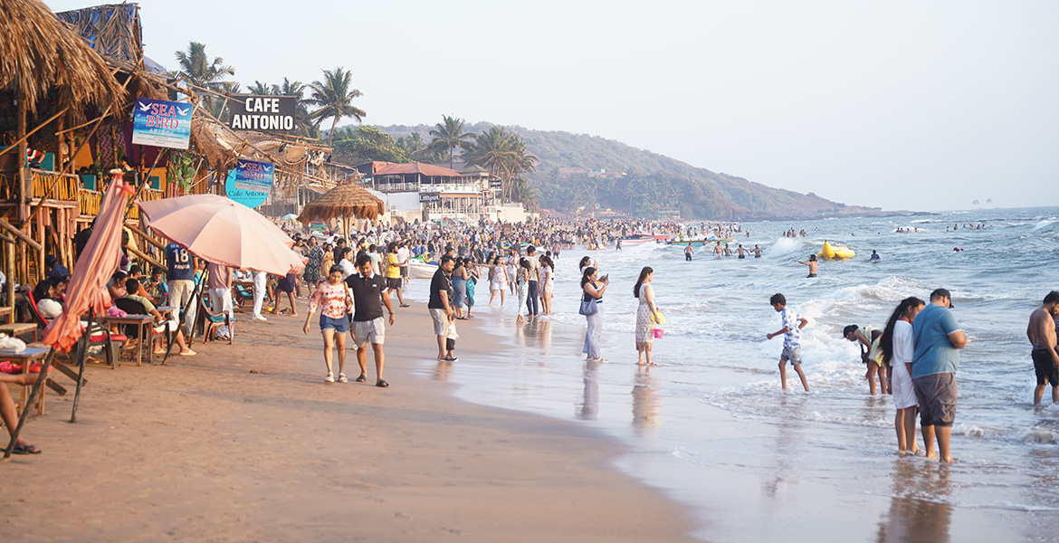 Crowded Anjuna Beach in Goa with tourists, beachside cafes, and sea waves during daytime