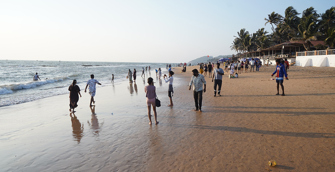 Visitors walking along Anjuna Beach in Goa with sea waves, palm trees, and sunset light