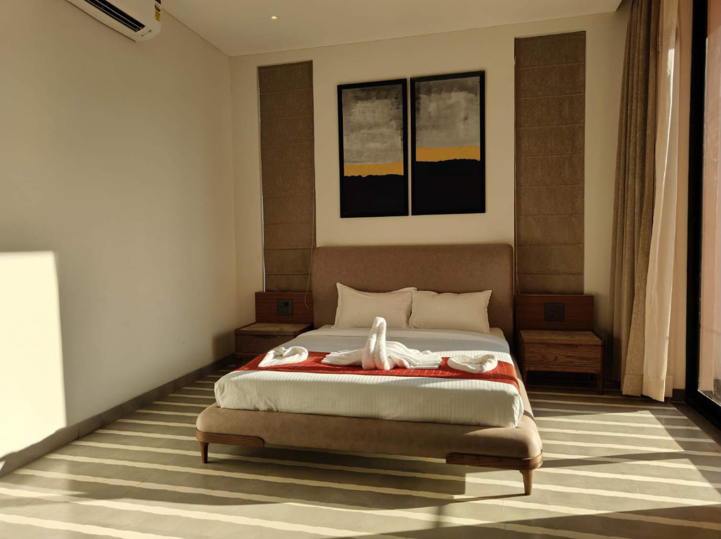 Brightly lit bedroom with strong floor shadows, a beige bed, and dark abstract art.