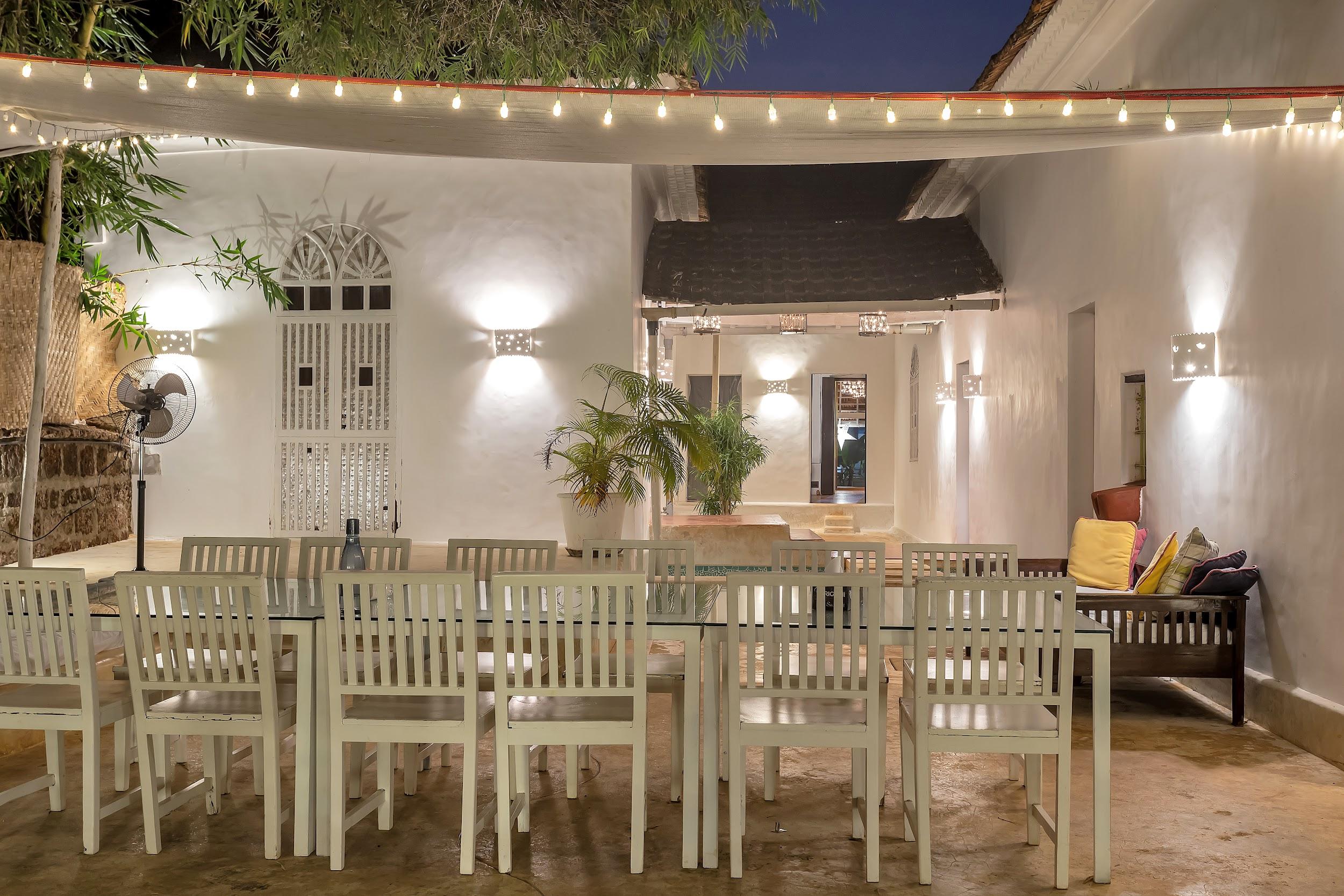 Night shot of the covered outdoor dining area with a large glass table, white chairs, and wall sconces